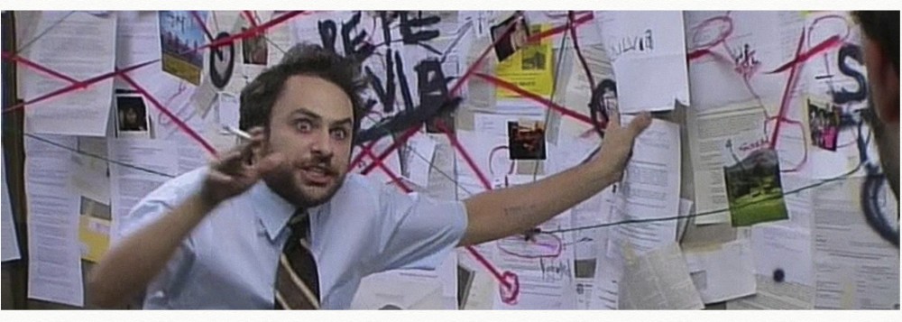 a wild-eyed man stands in front of a cork board covered with various documents with pins connected by red yarn